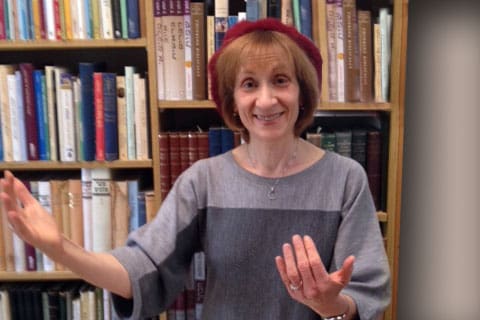 Judy Klitsner giving a welcoming gesture, standing in her office with a bookcase of books behind her.